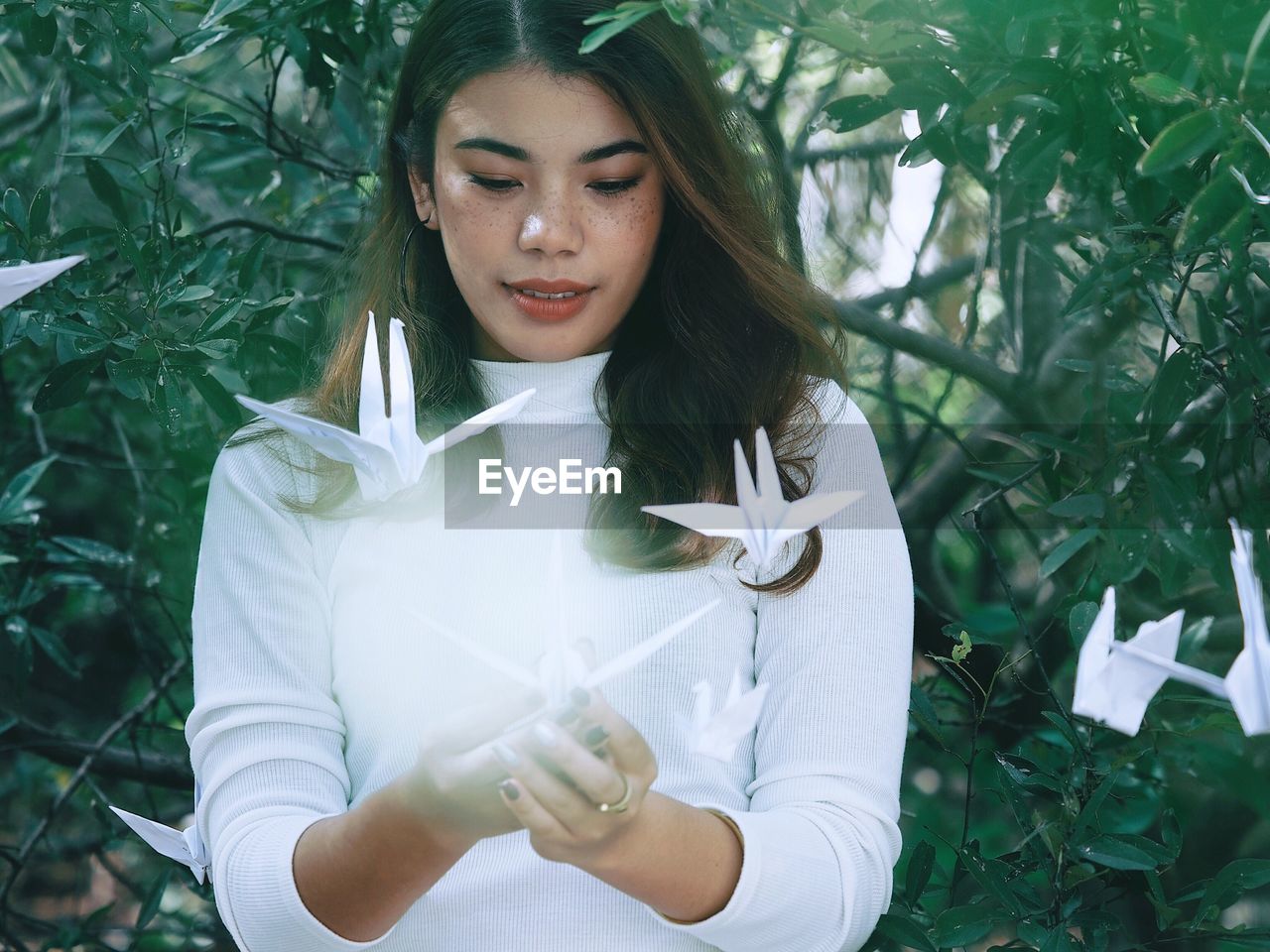 Beautiful young woman hanging flying paper cranes while standing against plants at park