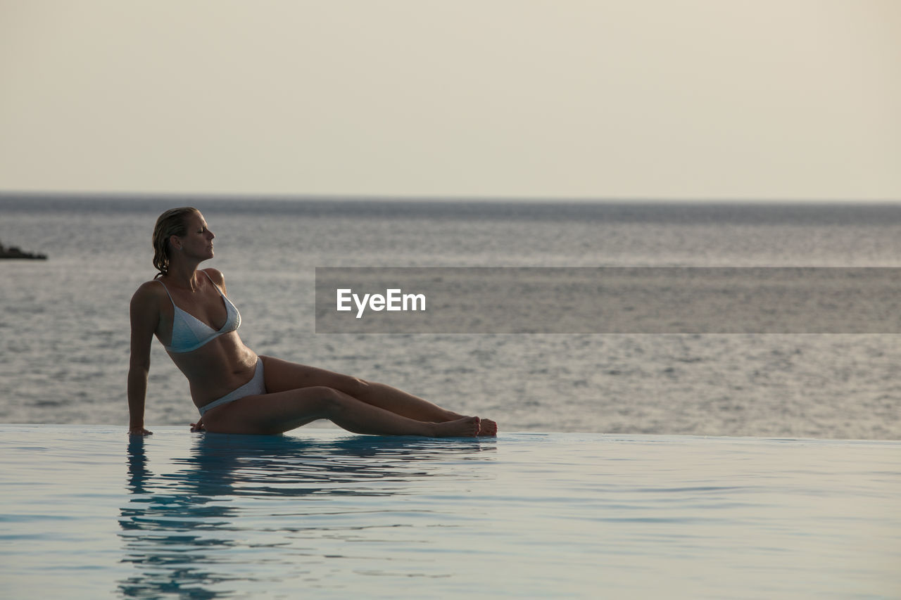 Full length of woman sitting by infinity pool against sea