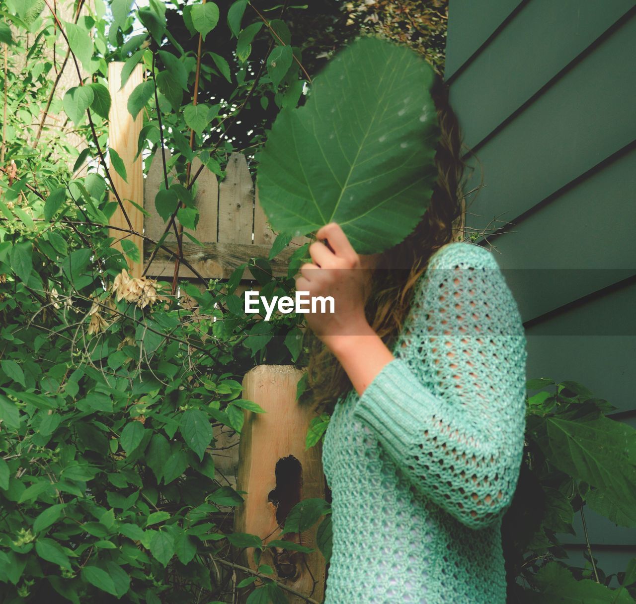 Side view of young woman hiding face with leaf in back yard