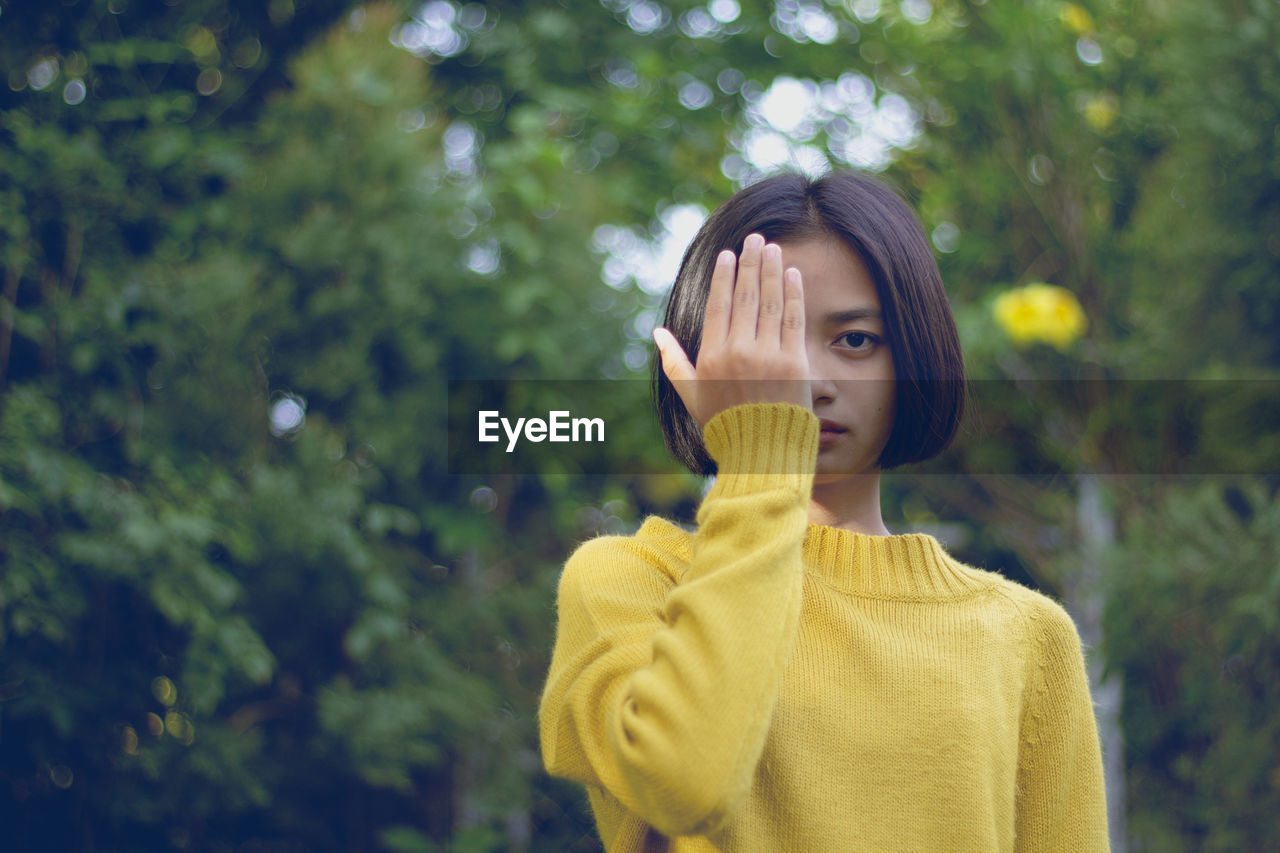 Portrait of teenage girl covering eye with hand against trees