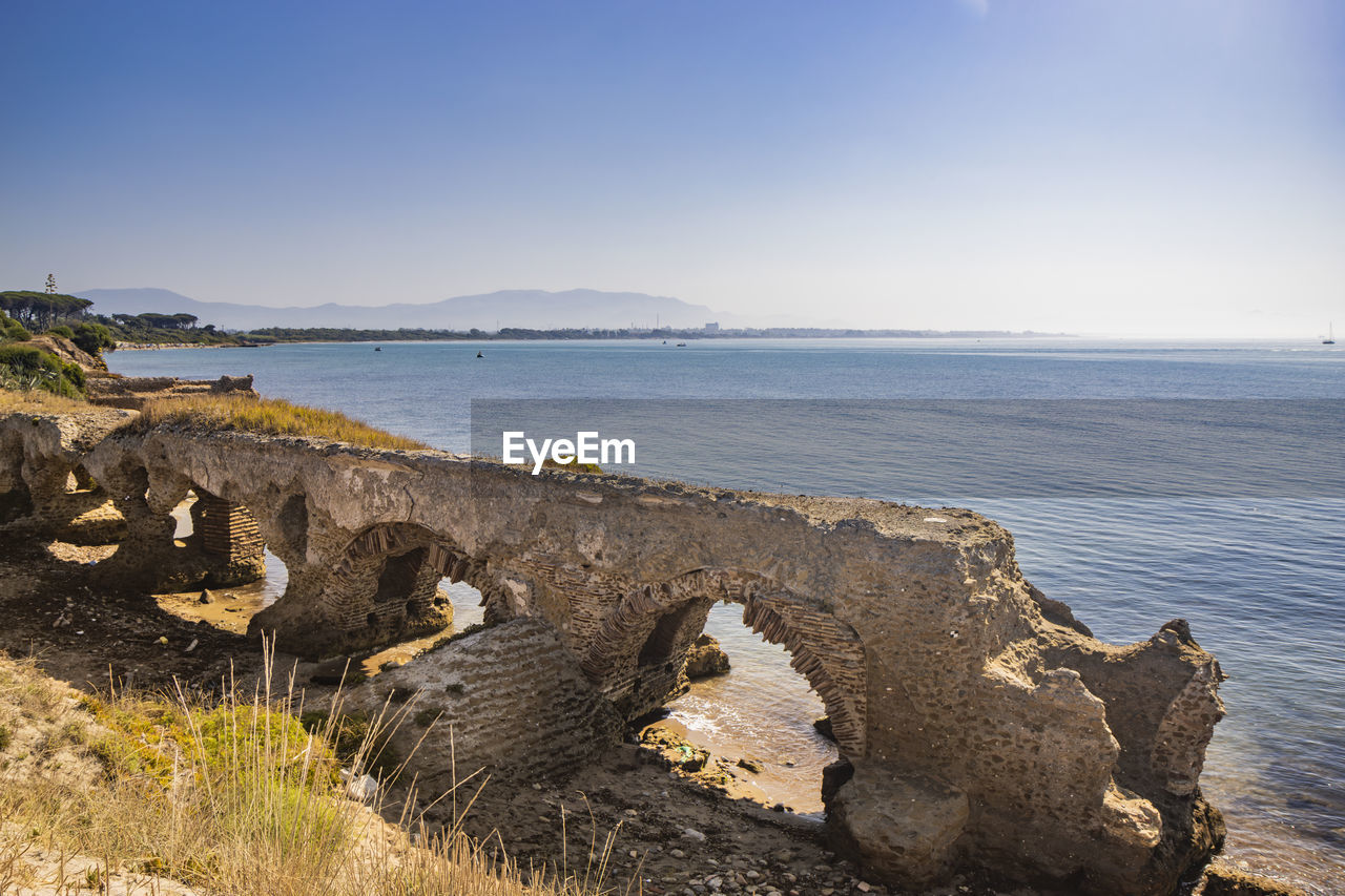 water, sea, sky, land, coast, shore, rock, beach, ocean, nature, scenics - nature, cliff, body of water, beauty in nature, travel destinations, wave, travel, terrain, horizon, environment, tranquility, coastline, landscape, no people, bay, horizon over water, tranquil scene, clear sky, architecture, outdoors, tourism, history, day, blue, sunny, sand, sunlight, cloud, seascape, the past, trip, holiday, non-urban scene, reflection, vacation