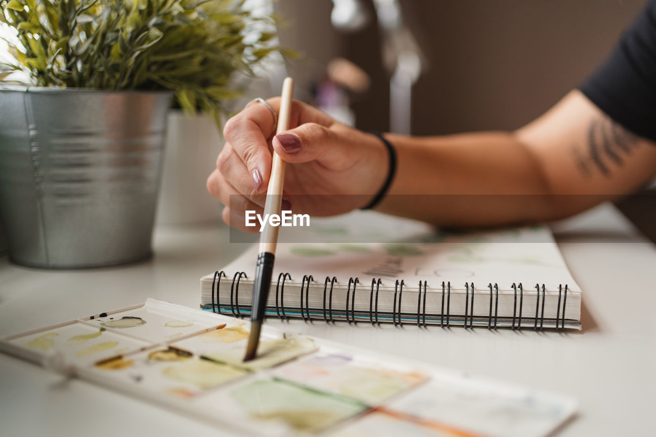 Closeup of unrecognizable woman using palette and watercolors while creating picture and sitting at table at home in paris