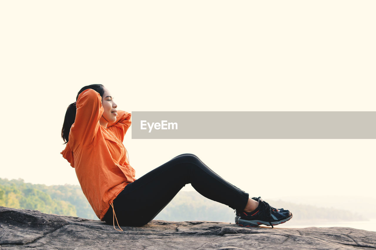 Young woman exercising on cliff against clear sky