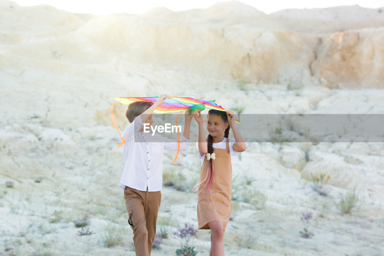 A girl and a boy carry bright colored kite over their heads while walking in mountains.