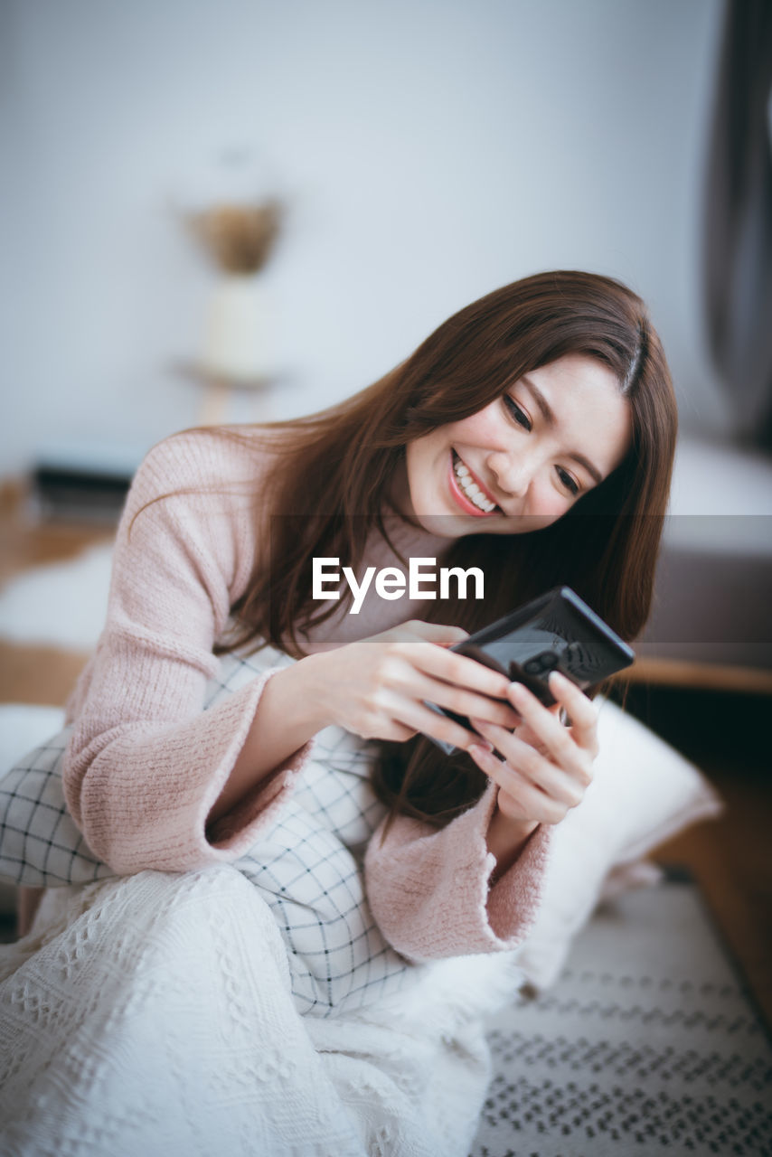 Smiling young woman using mobile phone while sitting on bed at home
