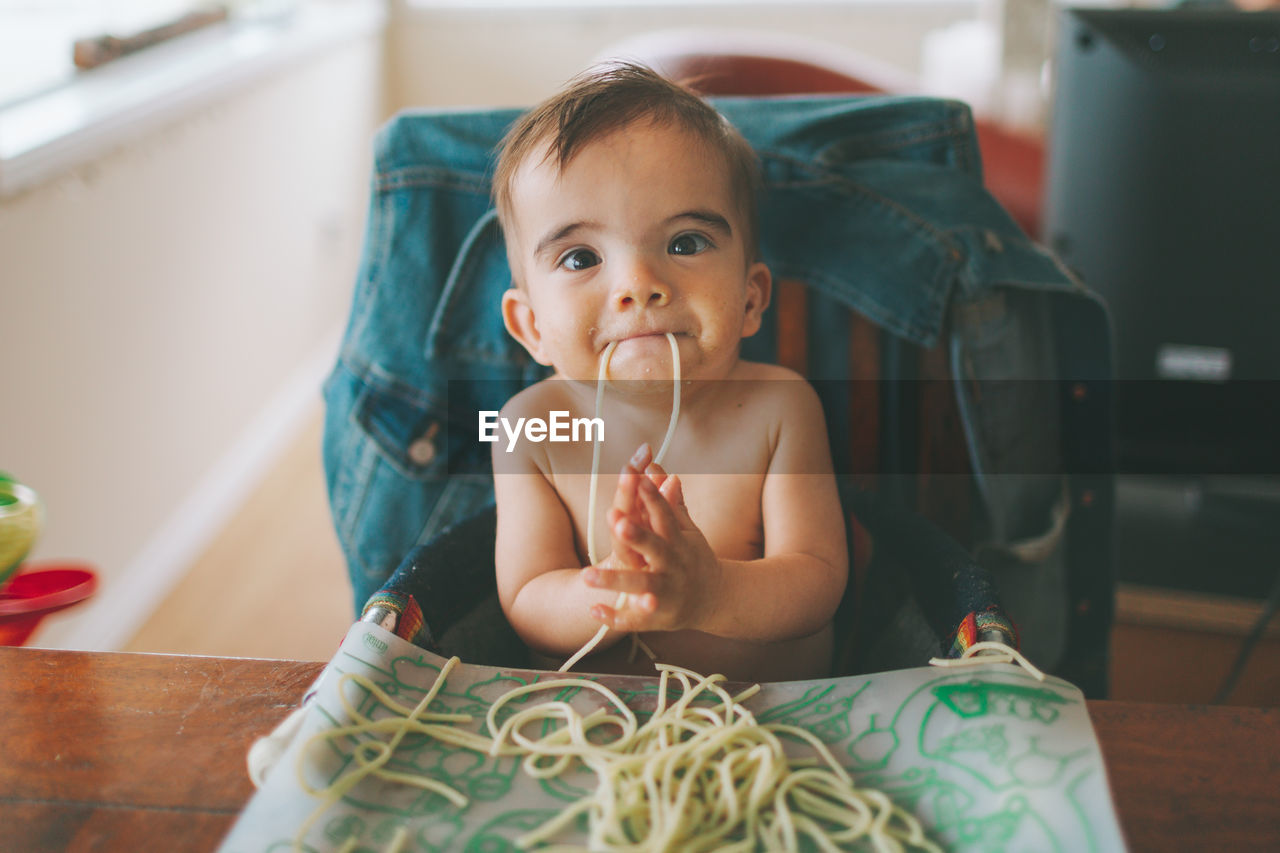 Portrait of shirtless baby boy eating spaghetti at home