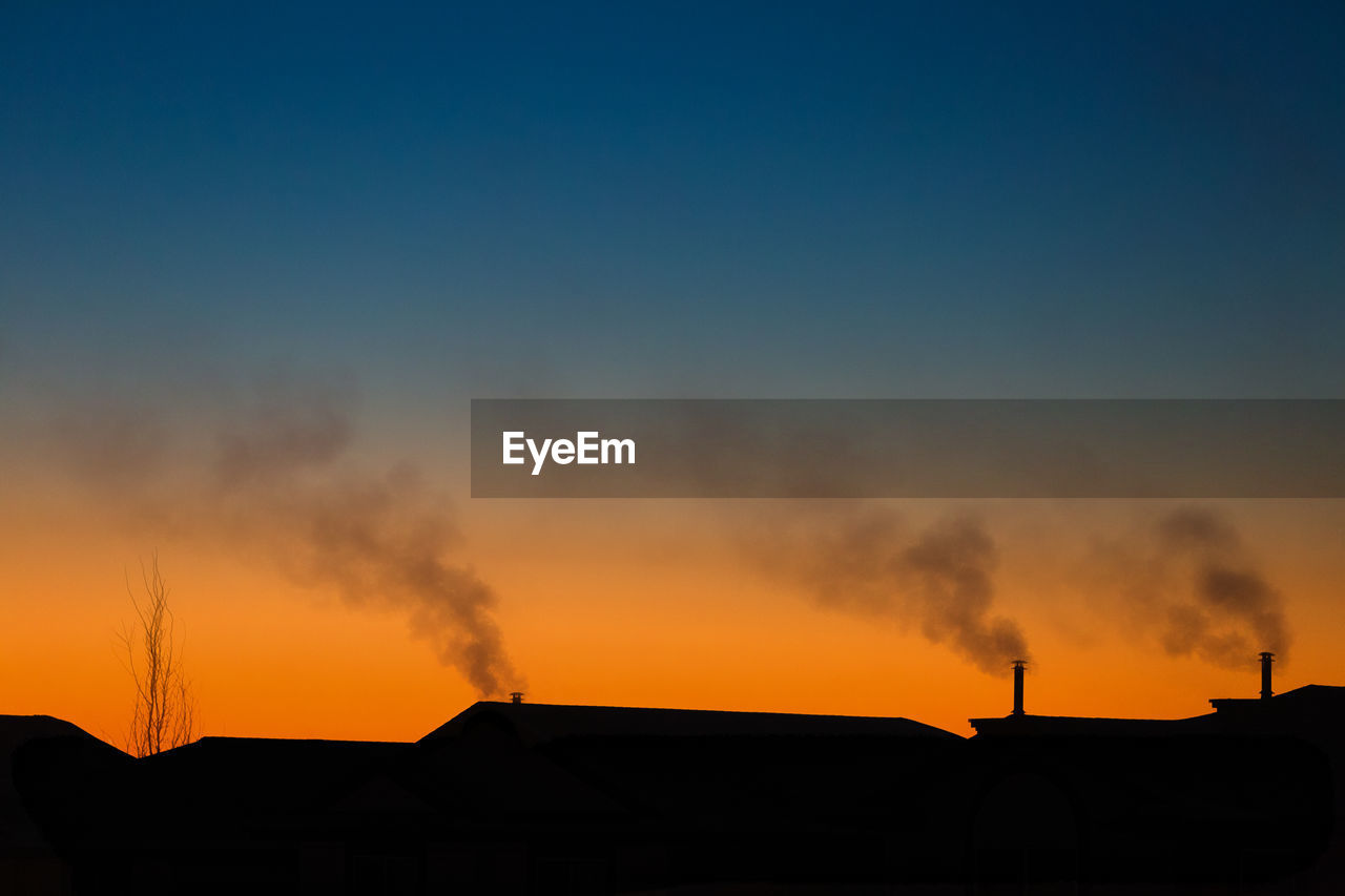 Smoke emitting from silhouette houses against clear sky during sunset