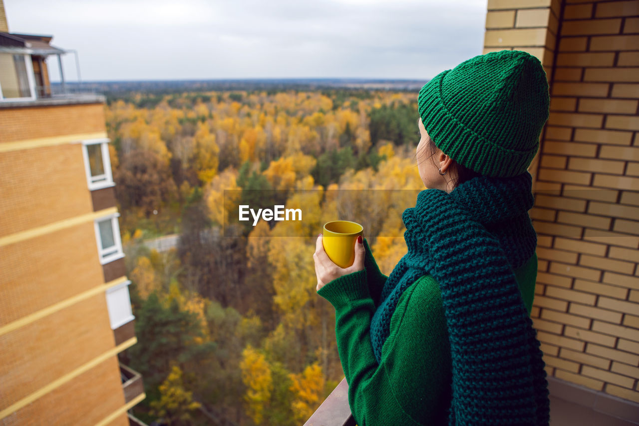 Woman in a green knitted sweater and a hat stands with a yellow mug on the balcony, autumn forest