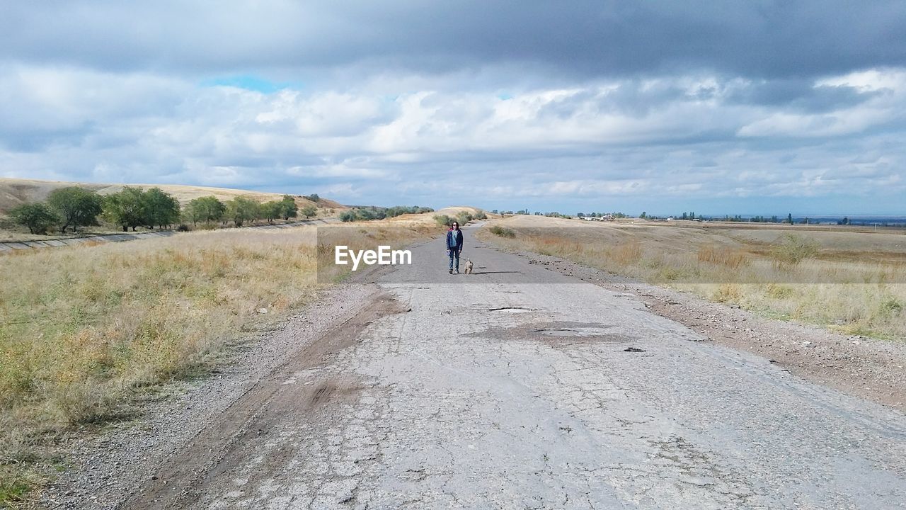 Distant view of woman standing with dog on road against cloudy sky