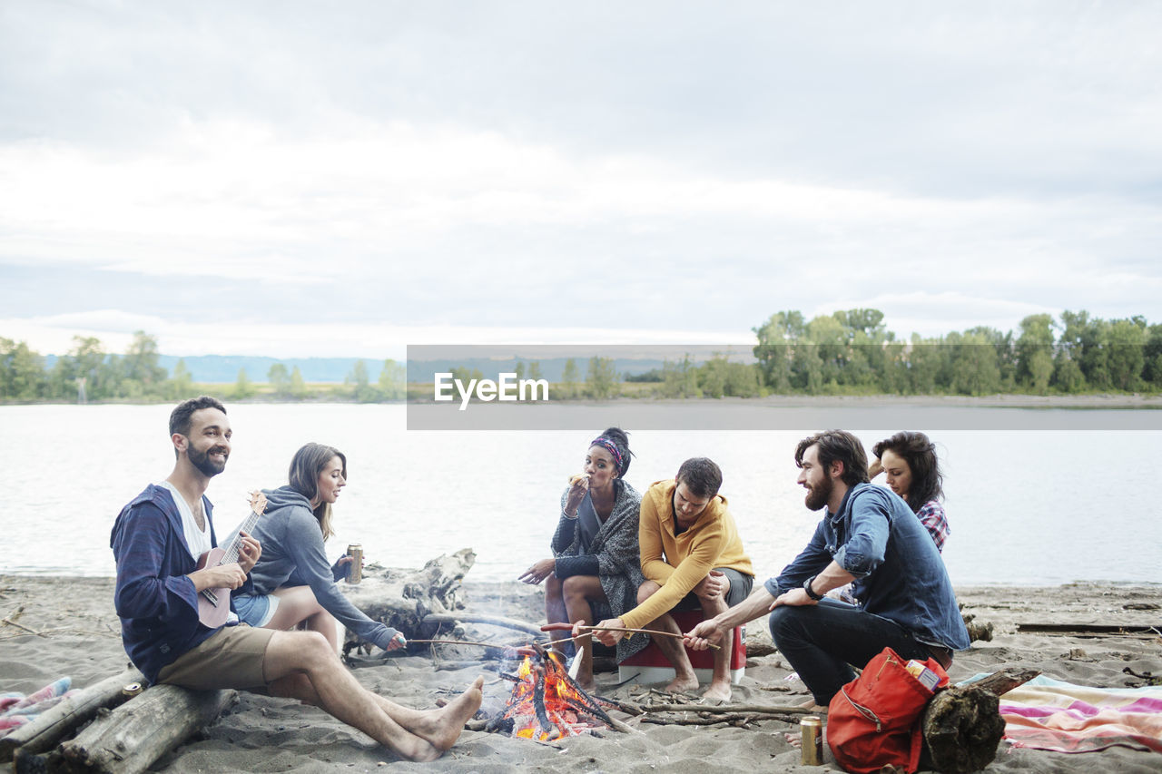 Happy friends roasting sausages on campfire at riverbank against sky