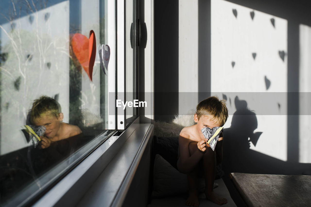 Shirtless boy making decorations while sitting by window at home