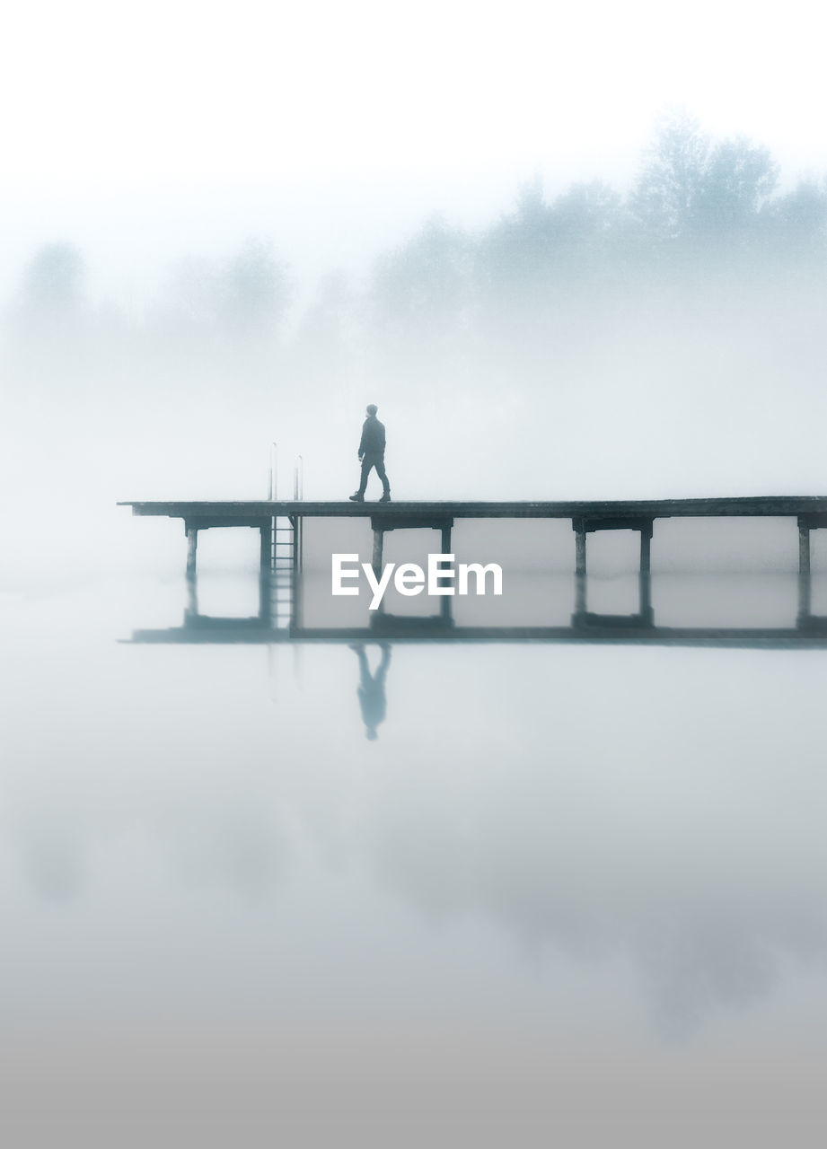 SILHOUETTE MAN STANDING ON LAKE AGAINST SKY DURING FOGGY WEATHER