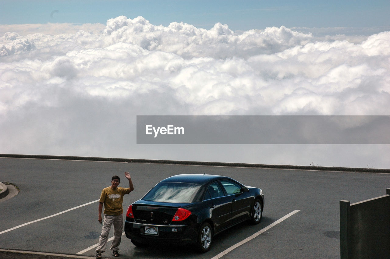 Man standing by car at parking lot against cloudy sky