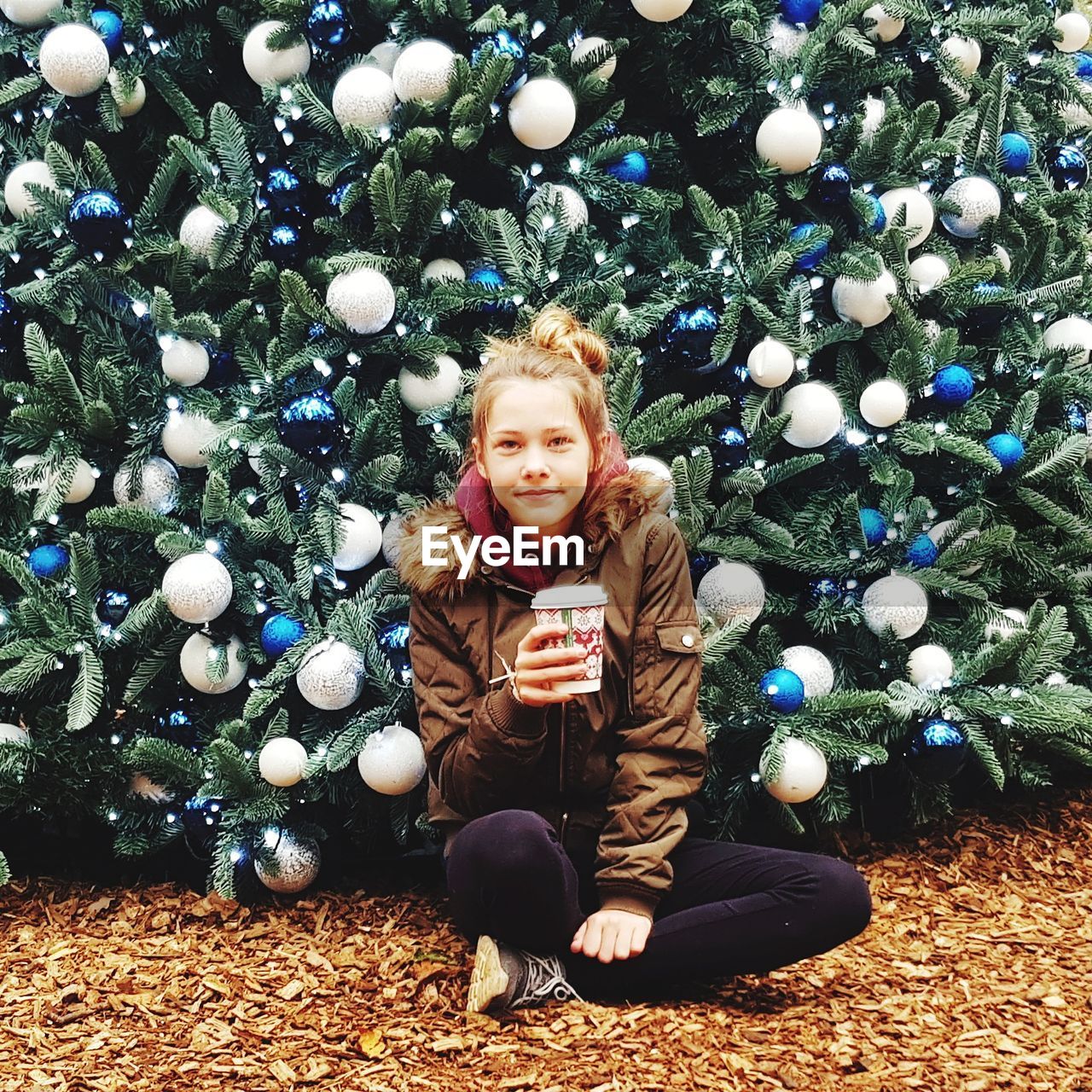 Portrait of girl holding drink while sitting on field against christmas tree