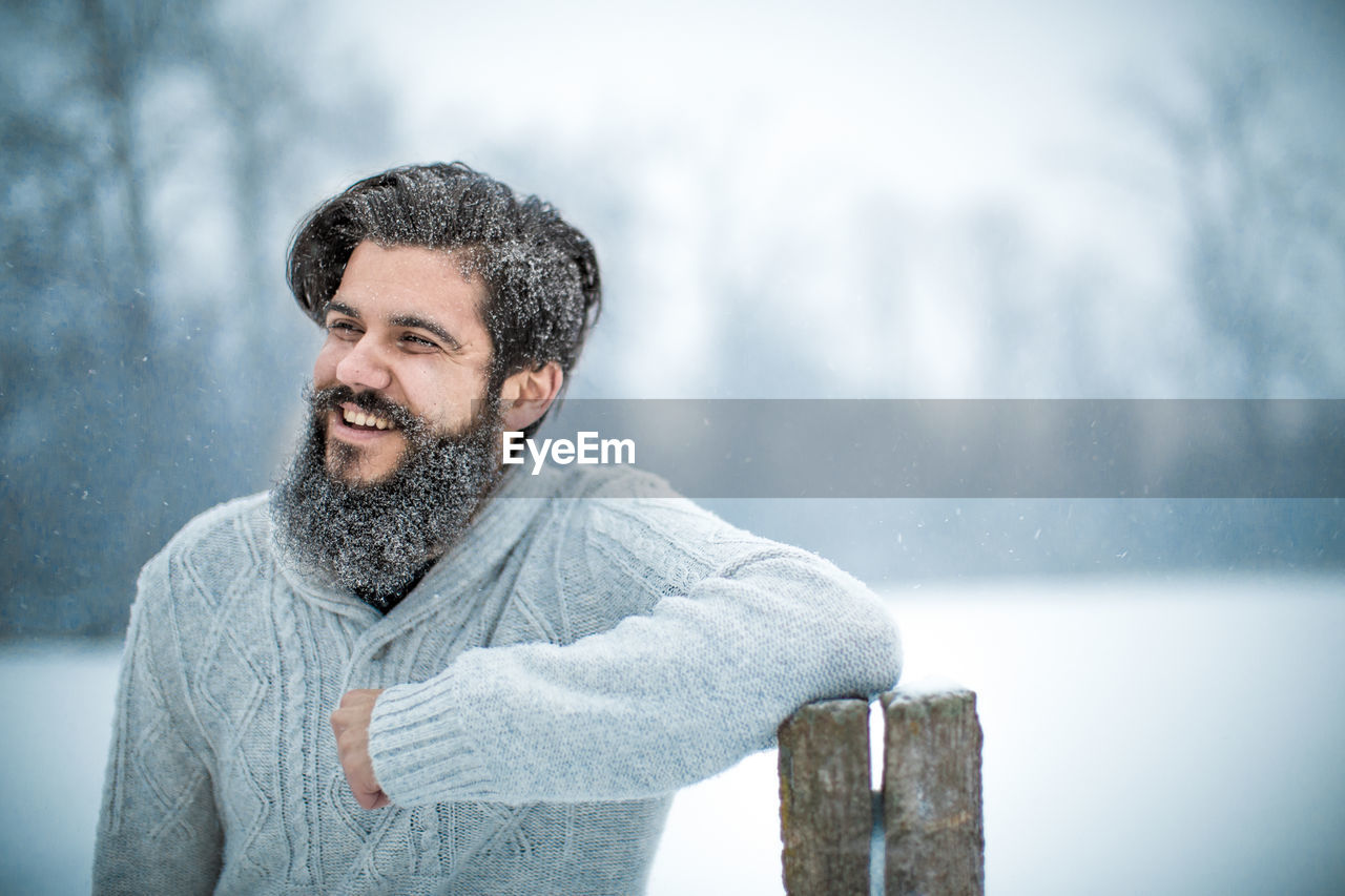 Happy young bearded man with arms crossed standing during snowfall