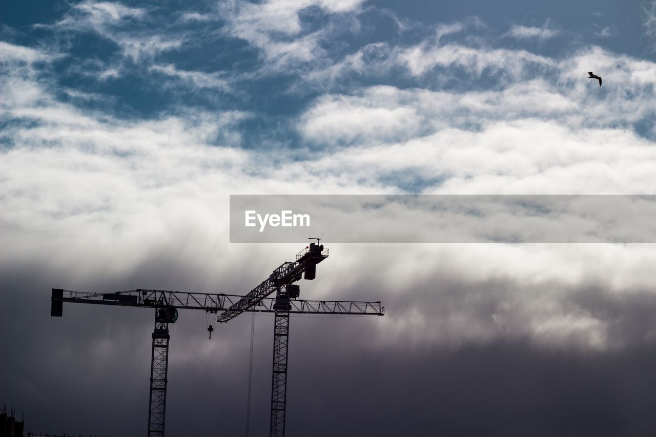 Silhouette cranes at construction site against cloudy sky