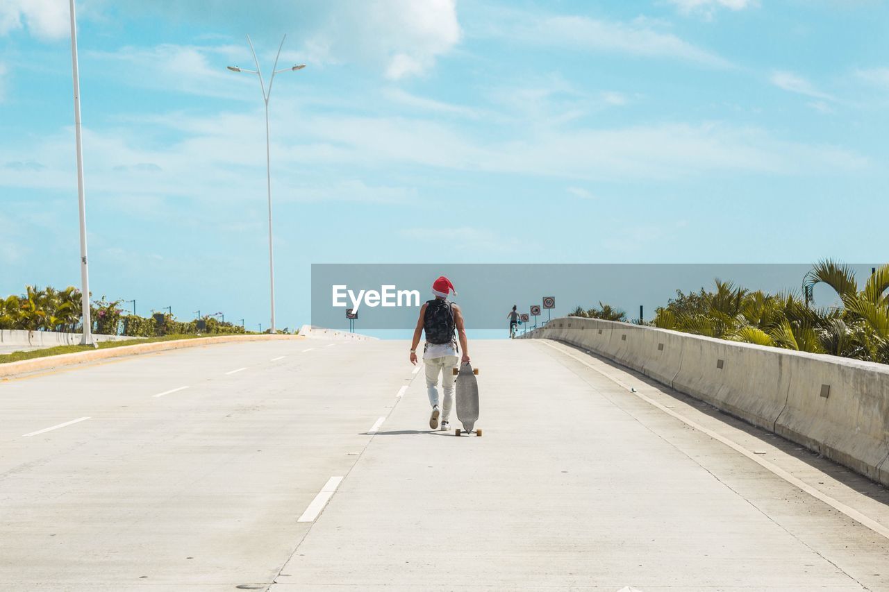 Rear view of man with skateboard walking on road against sky