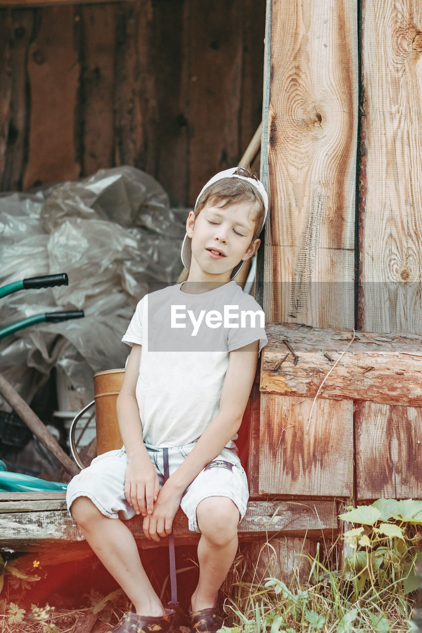 A tired boy sits leaning against the barn door with his eyes closed. 