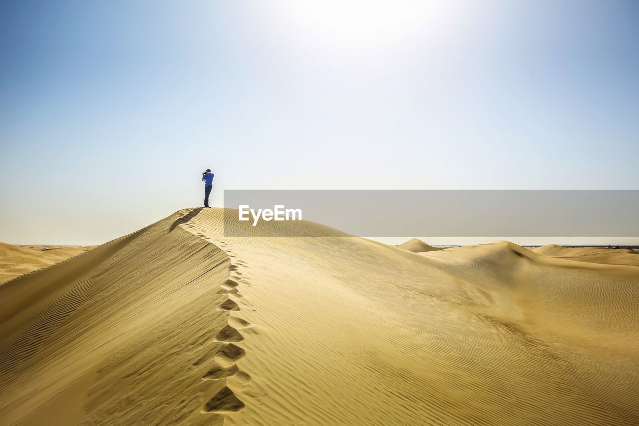 Boy photographing while standing on sand dune in desert against sky