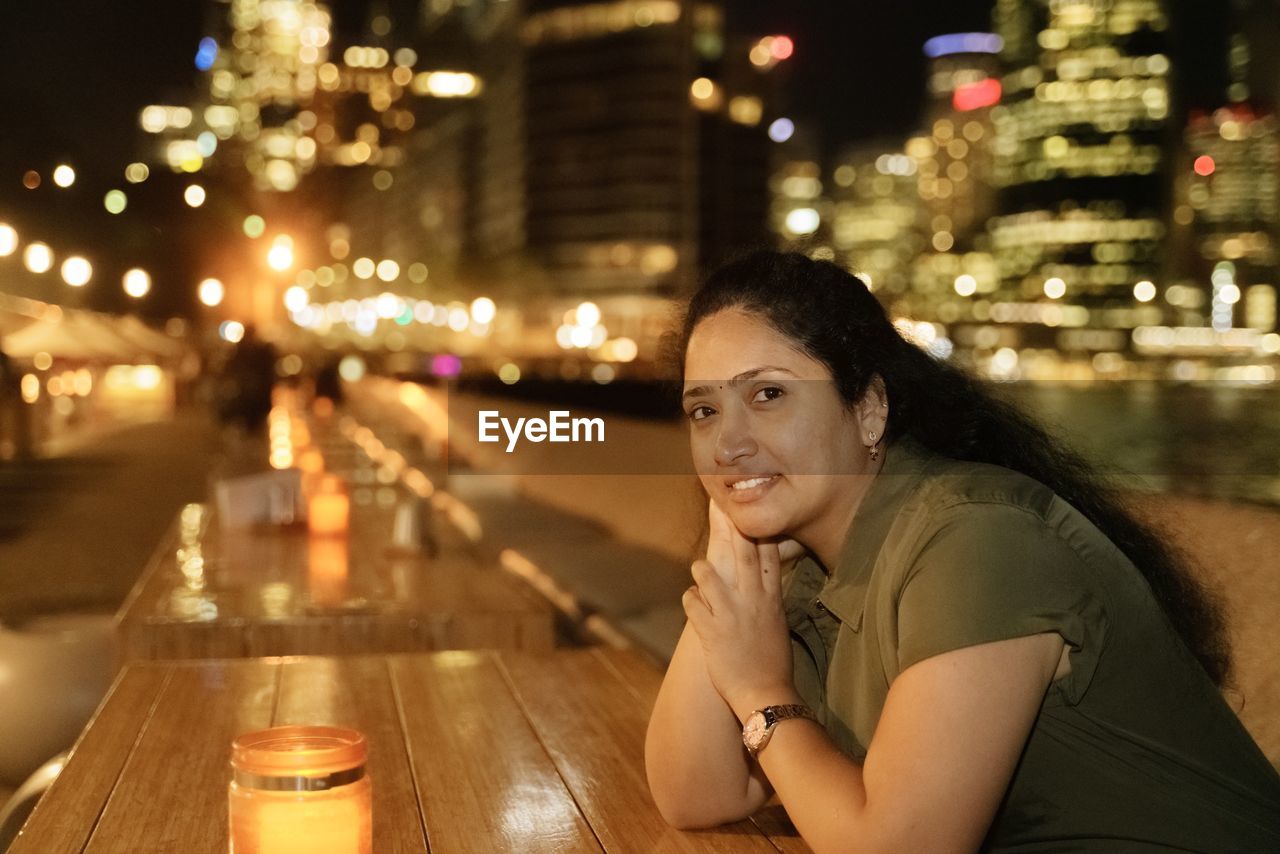 PORTRAIT OF WOMAN SITTING AT ILLUMINATED CITY AT NIGHT