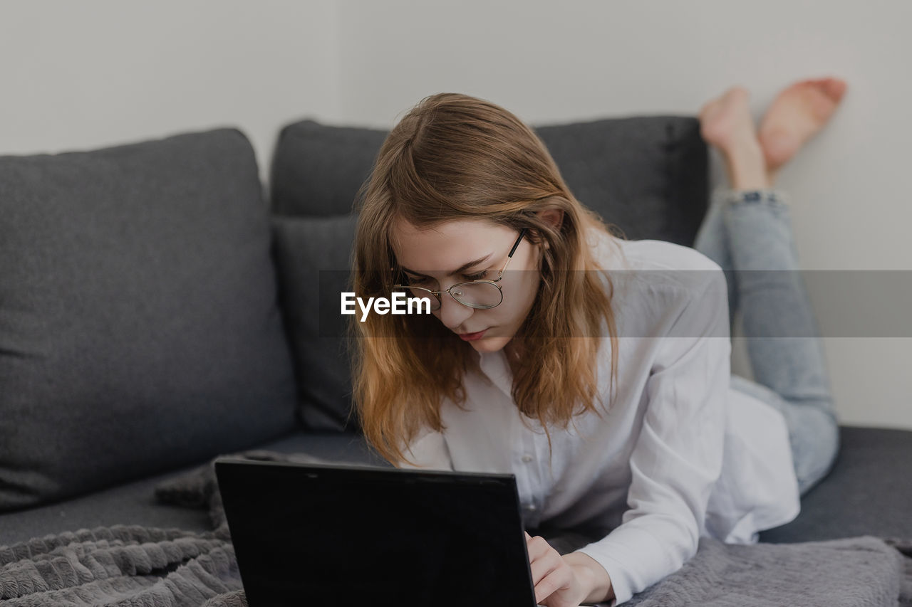 Attractive young woman working from home - female entrepreneur sitting on sofa with laptop computer and checking cell phone from comfort of home