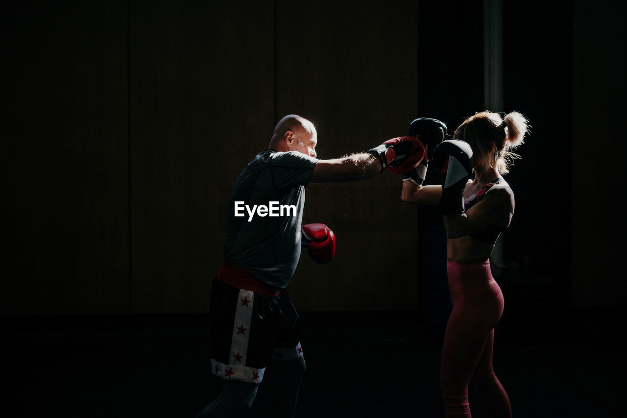 Side view of concentrated young sportswoman athlete in boxing gloves training fighting with professional male coach in modern gym