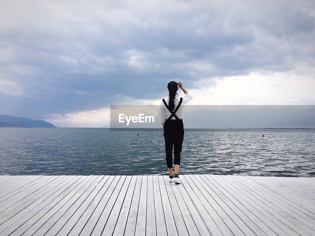 Full length rear view of woman in suspenders standing on pier against sea and cloudy sky