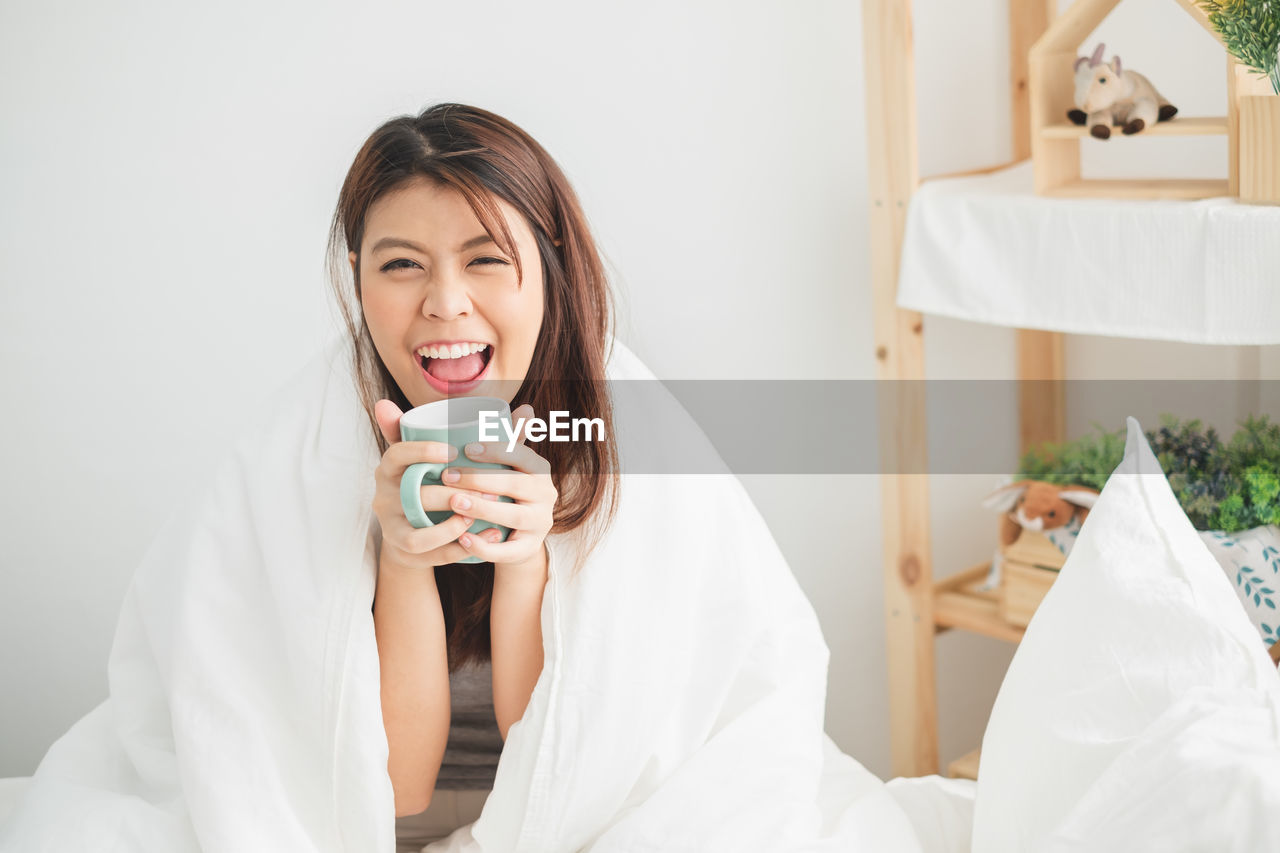 Portrait of young woman holding coffee cup sitting on bed at home