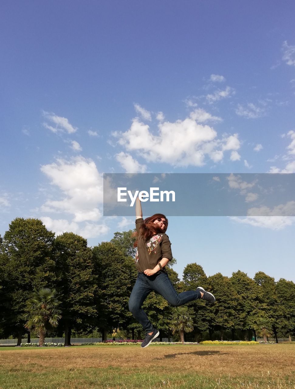 Happy mature woman with arms raised jumping on field against sky