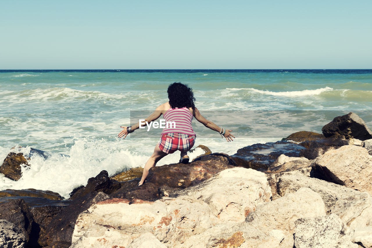 Rear view of woman with arms outstretched crouching on rock at beach against clear sky