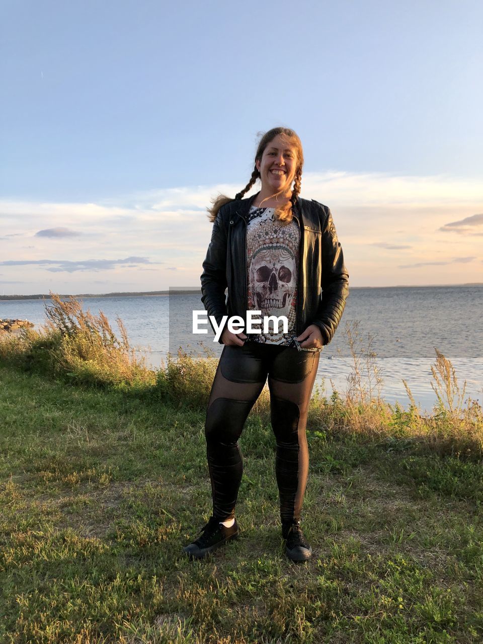 sky, one person, sea, full length, land, water, nature, standing, beach, front view, adult, portrait, beauty in nature, grass, leisure activity, casual clothing, smiling, looking at camera, young adult, women, day, lifestyles, scenics - nature, cloud, plant, horizon, clothing, horizon over water, outdoors, holiday, happiness, emotion, sunlight, trip, vacation, copy space, tranquility, travel, coast, landscape