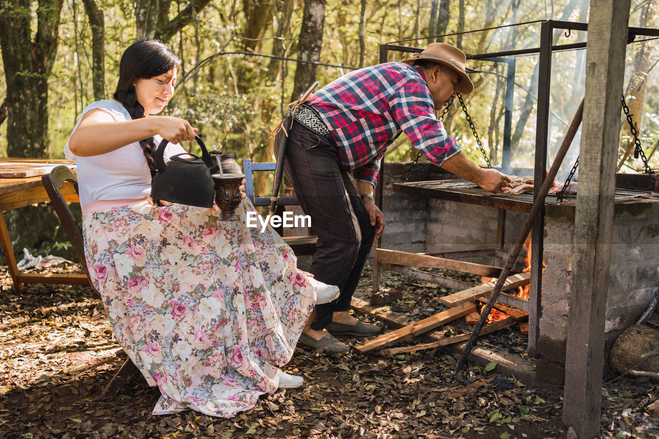 Smiling argentinian female pouring hot water from kettle into calabash gourd against male partner grilling meat above fire in campsite