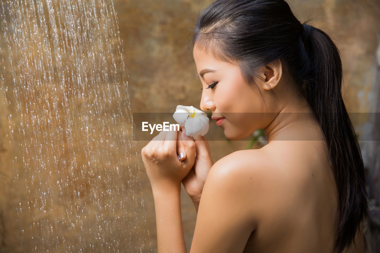 Rear view of topless young woman holding flower while taking bath in shower