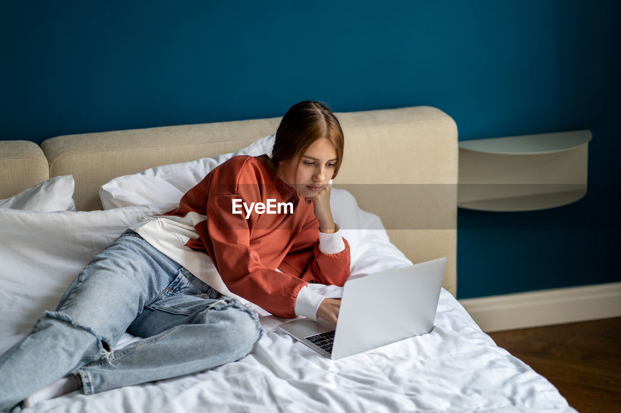 Thoughtful interested teen girl student lying on bed use laptop college study online webinar at home