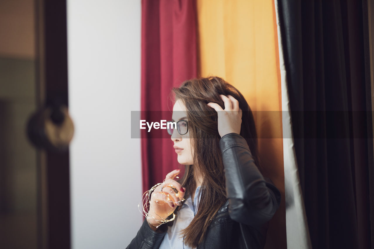 Young woman with hand in hair looking away while standing against curtains at home