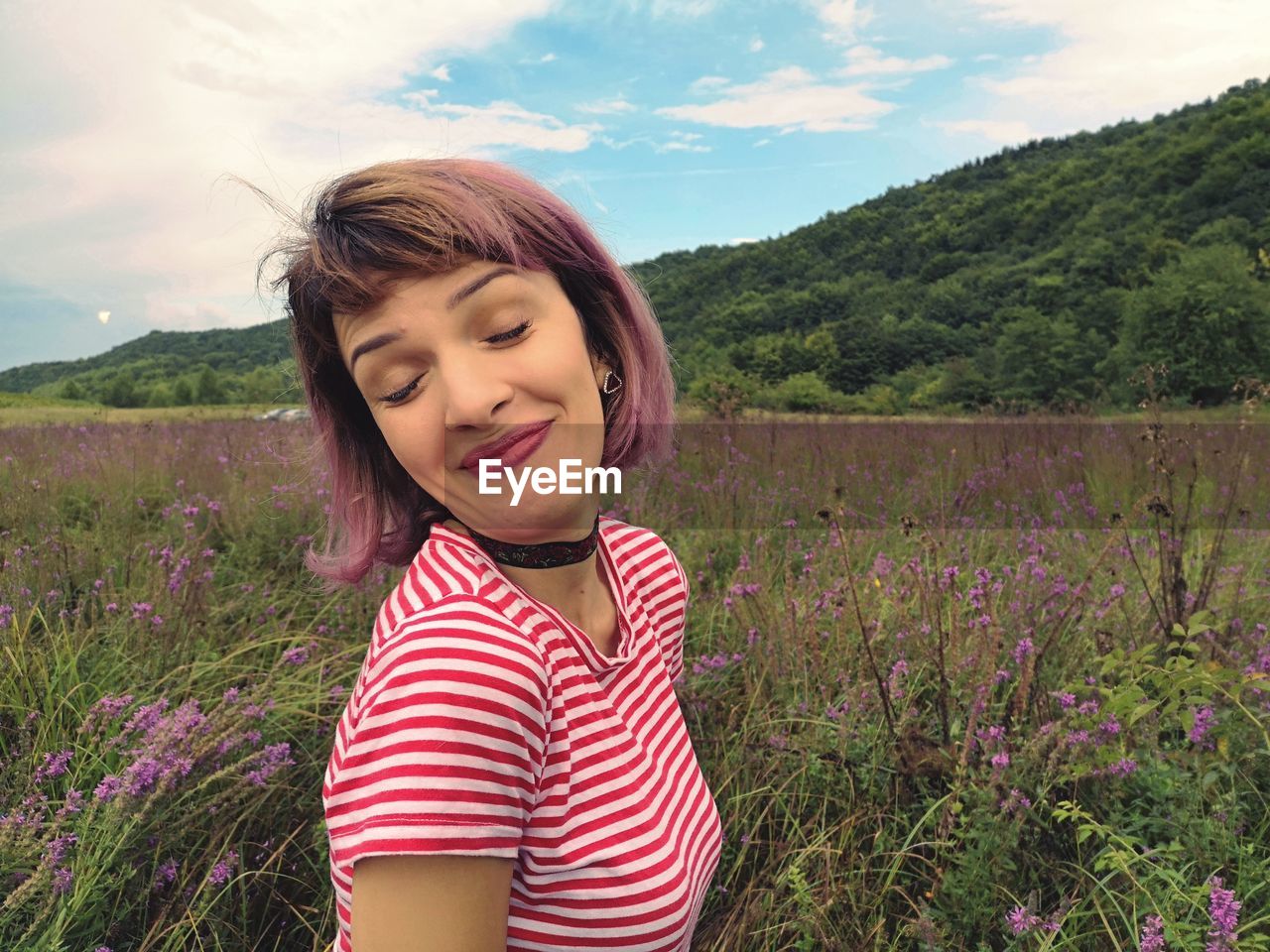 Smiling woman with eyes closed standing by plants on field against cloudy sky