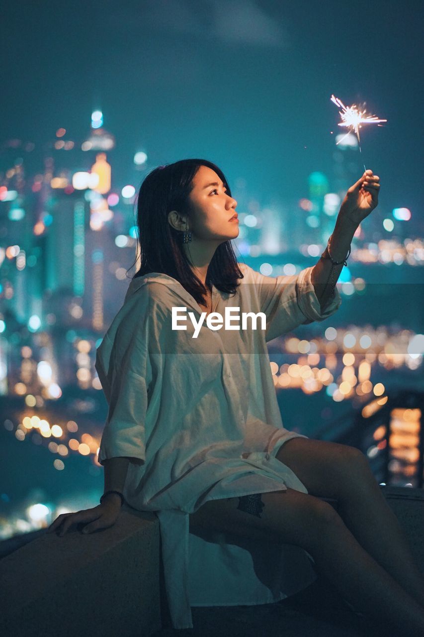 Young woman holding sparkler in city against sky at night