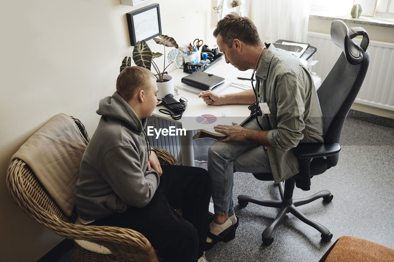 High angle view of school male discussing over chart with nurse student sitting at table in office