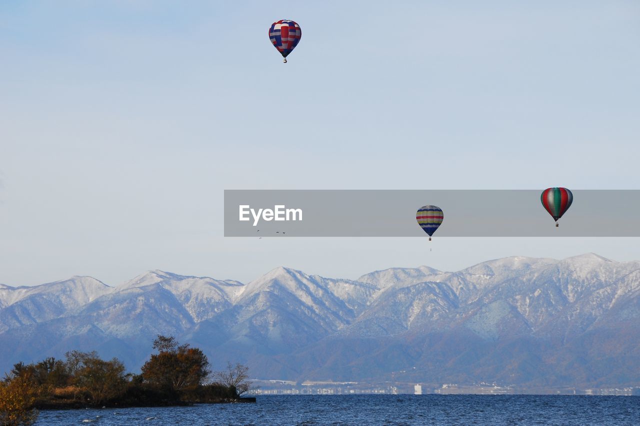 Hot air balloon flying over lake against clear sky and snowy mountains