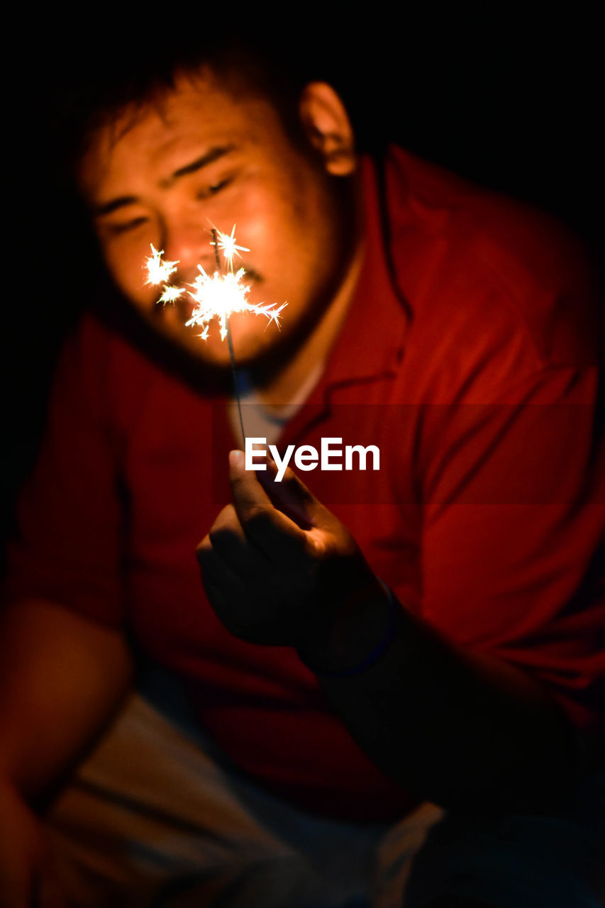 Smiling overweight man holding illuminated sparkler while sitting at night