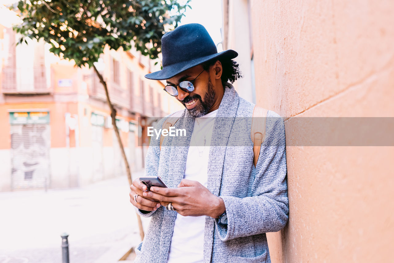 Hispanic male in stylish clothes with hat and sunglasses leaning on wall and browsing cellphone while standing on pavement on city street