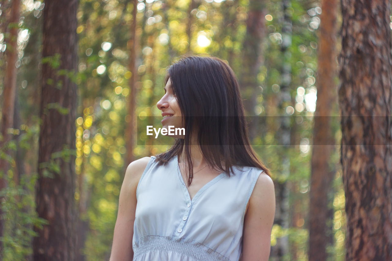 Woman looking away while standing against trees in forest