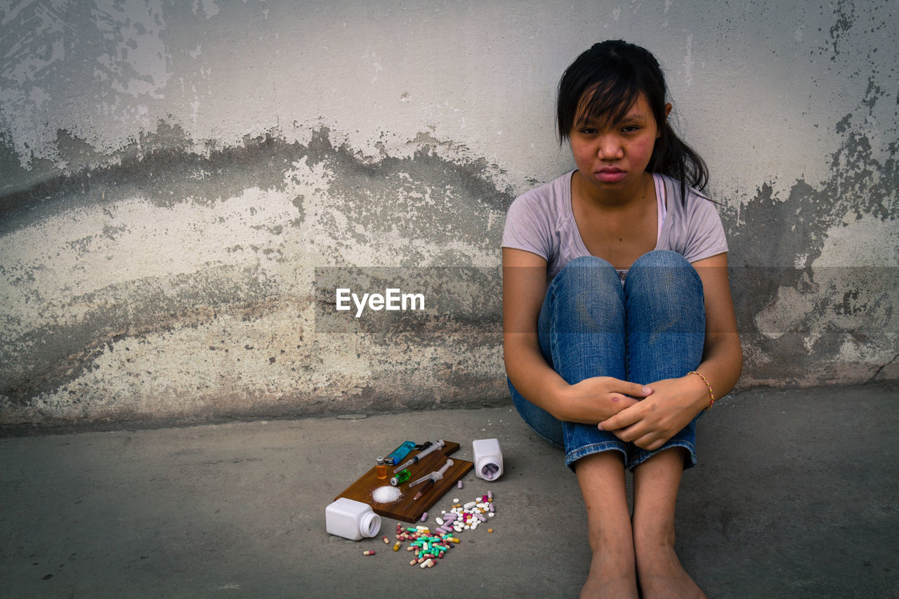 Depressed teenage girl sitting by narcotics against wall