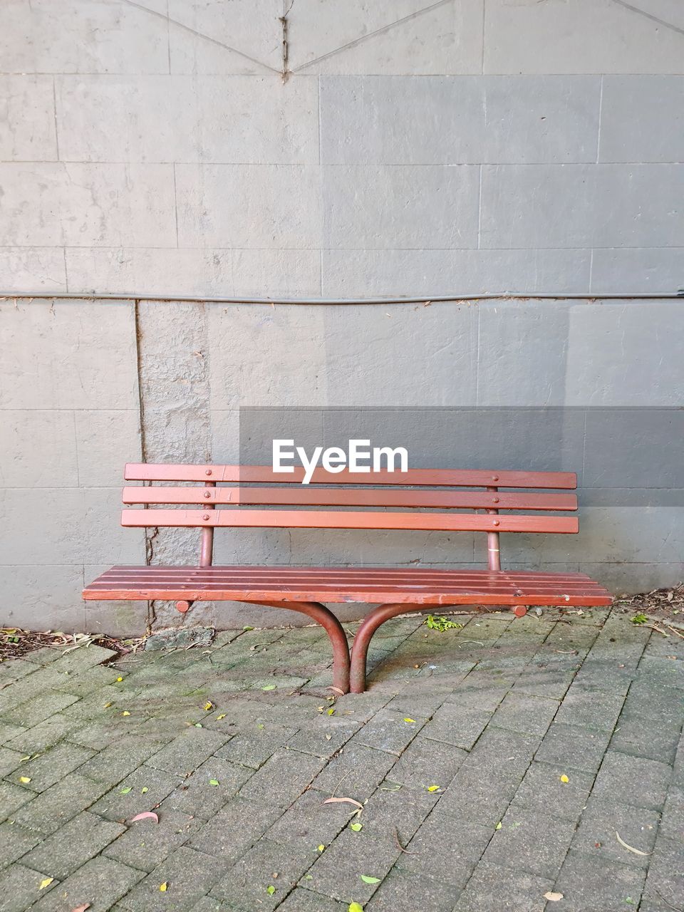 Empty brown wooden bench on footpath against grey brick wall with tiled floor.