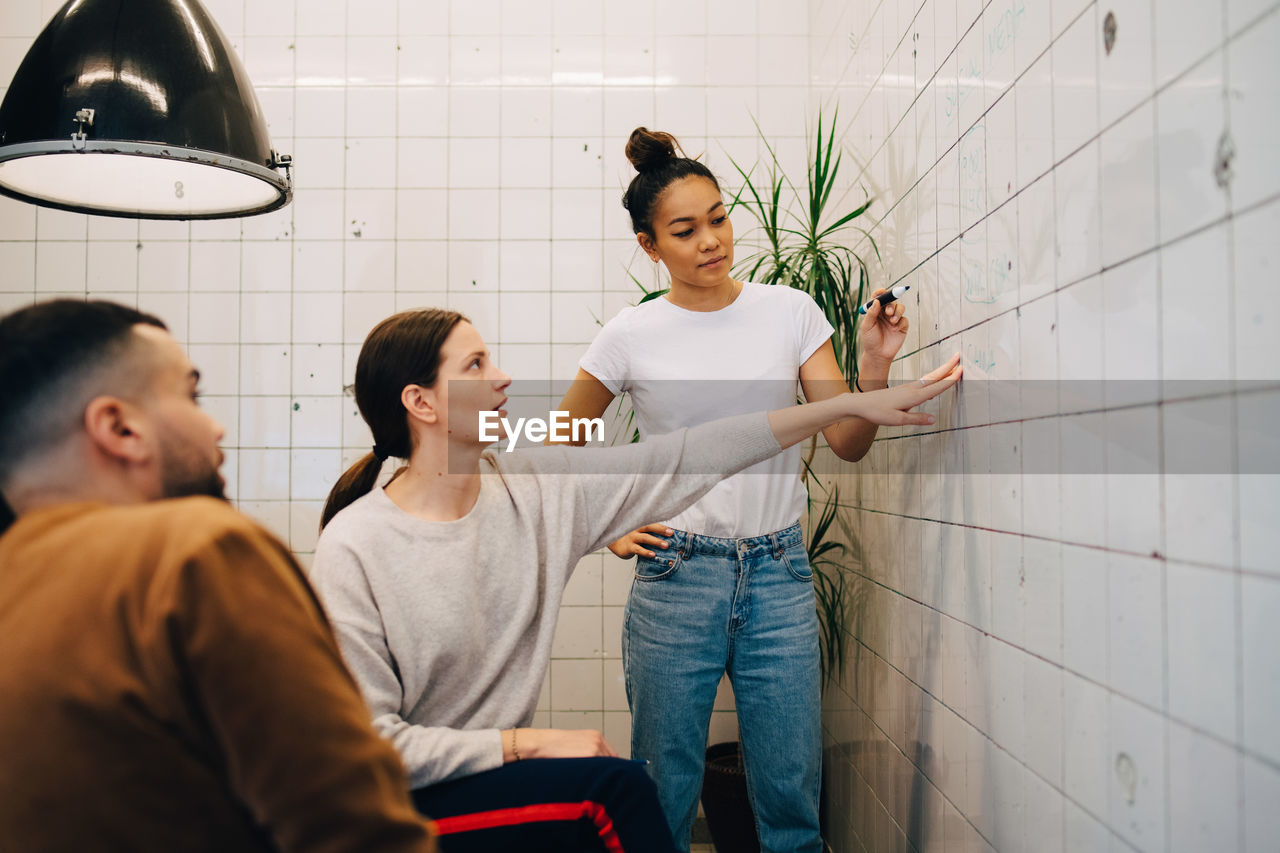 Young businesswoman brainstorming colleagues while writing strategy on tile wall at creative office
