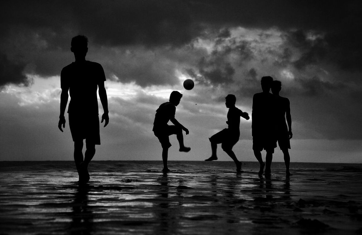 Silhouette boys playing soccer on beach against cloudy sky