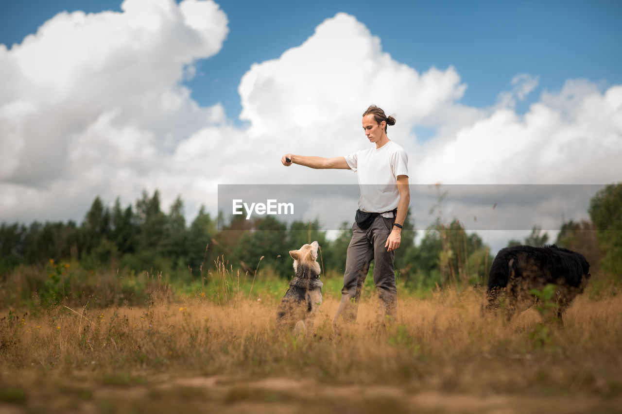 Man playing with dogs while standing on grassy land against cloudy sky