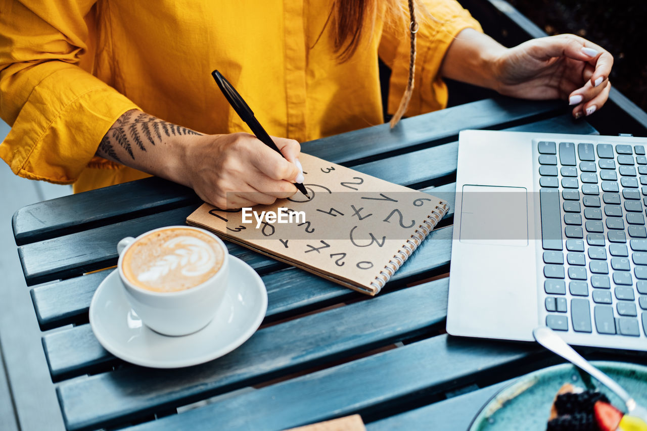 Woman freelancer writing numbers in notepad while working with laptop in street cafe. time