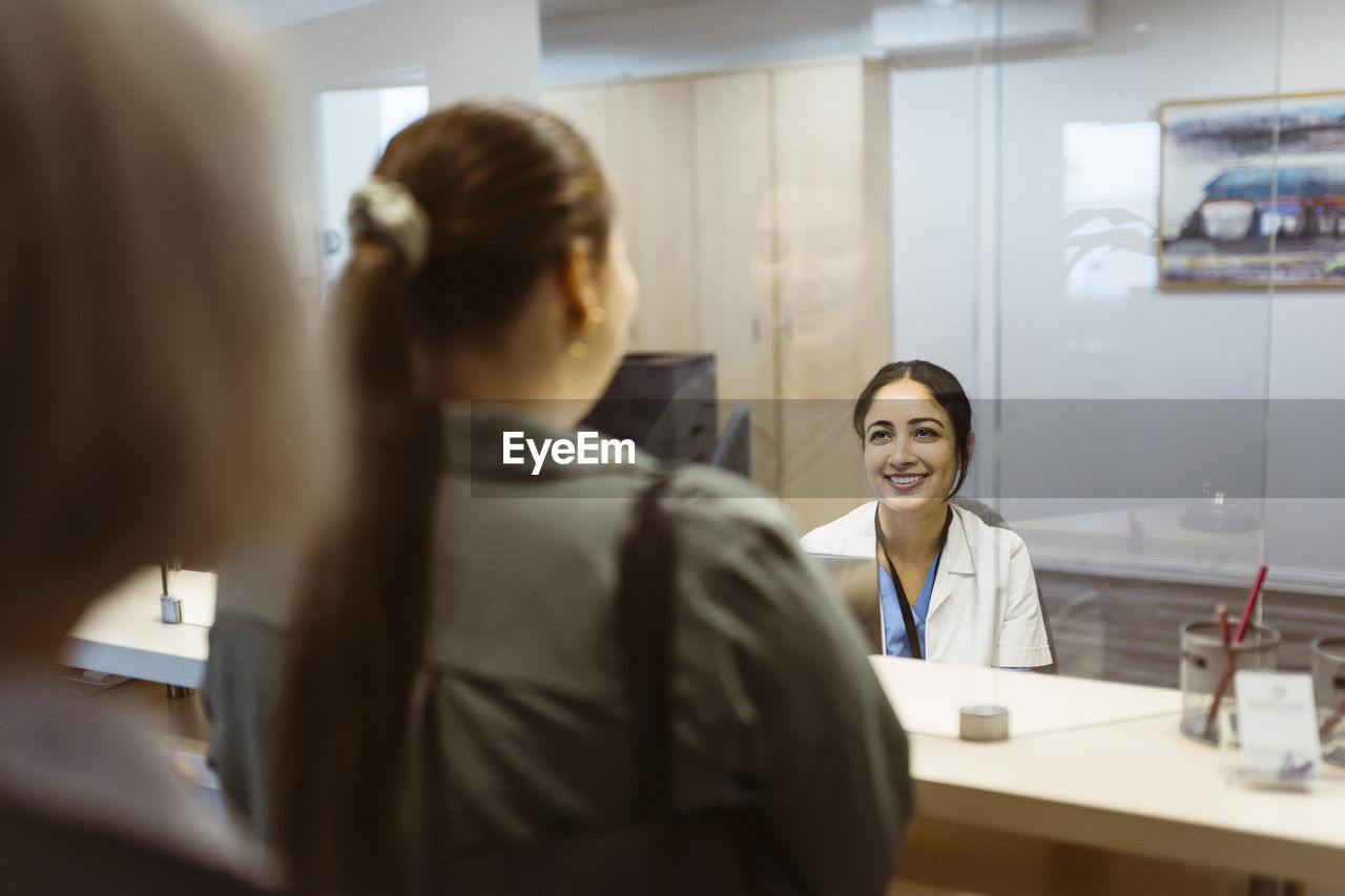 Smiling receptionist looking at woman through glass shield in hospital