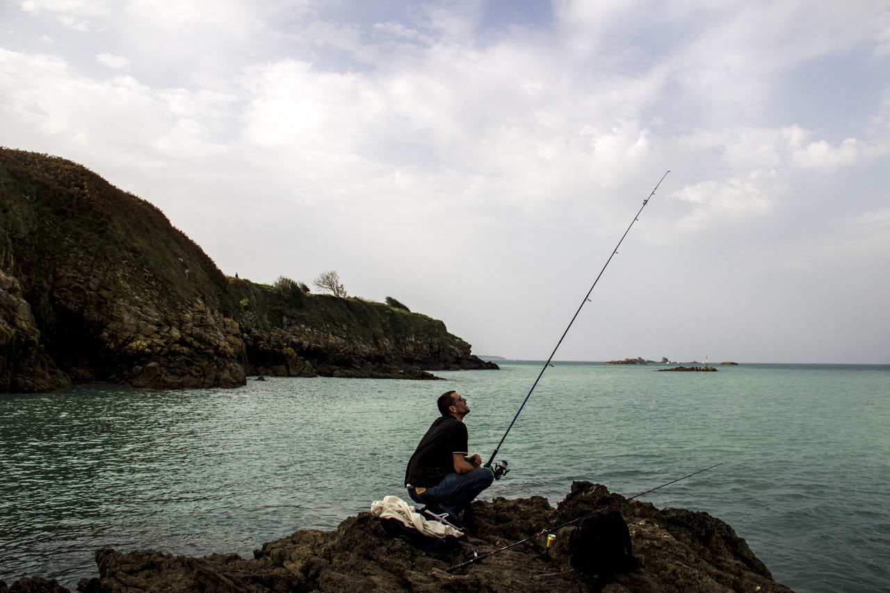 water, rock, sky, angling, fishing rod, rod, fishing, sea, nature, one person, activity, cloud, beauty in nature, casting, leisure activity, recreation, scenics - nature, coast, sitting, shore, fisherman, land, vacation, trip, holiday, adult, full length, men, beach, fly fishing, relaxation, tranquil scene, tranquility, bay, lifestyles, outdoors, idyllic, day, nautical vessel, travel, adventure, rock formation, ocean, person, non-urban scene, side view, sports, travel destinations